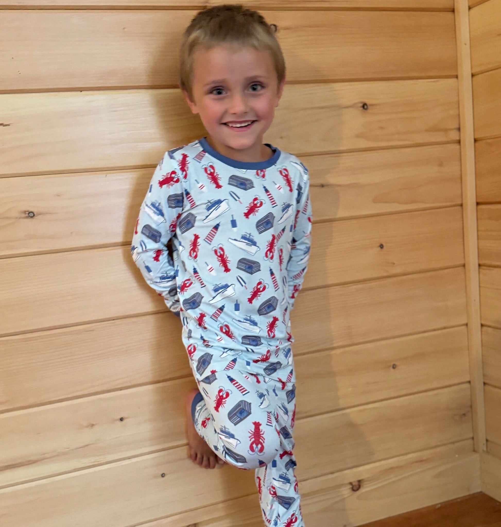 A toddler wearing a two-piece pajama set with a pattern of Maine lobster fishing inspired print with red lobsters, lobster boats, buoys, and lighthouses. standing against a wooden backdrop.